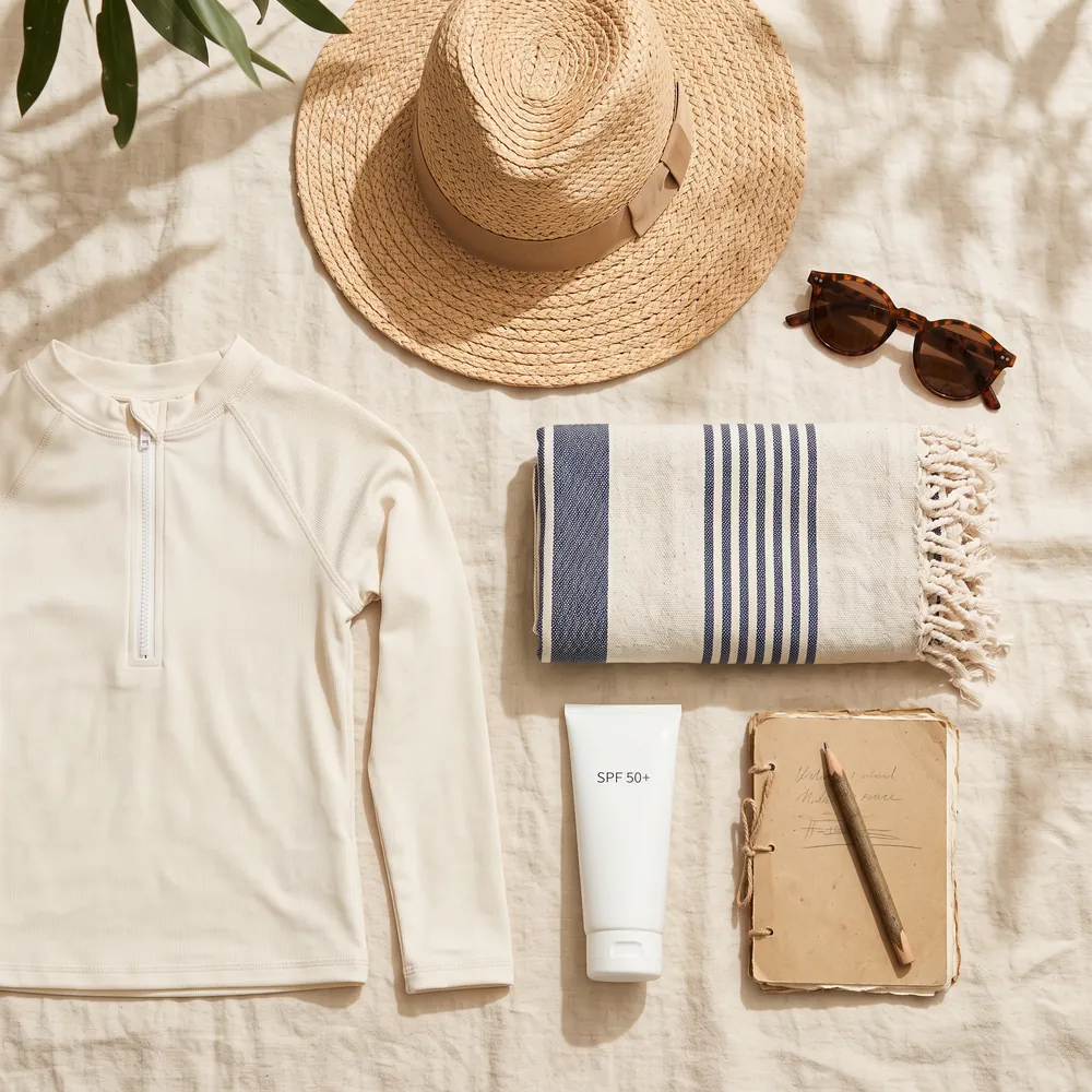 A sun hat, sunglasses, sunscreen and a notebook laid out on linen